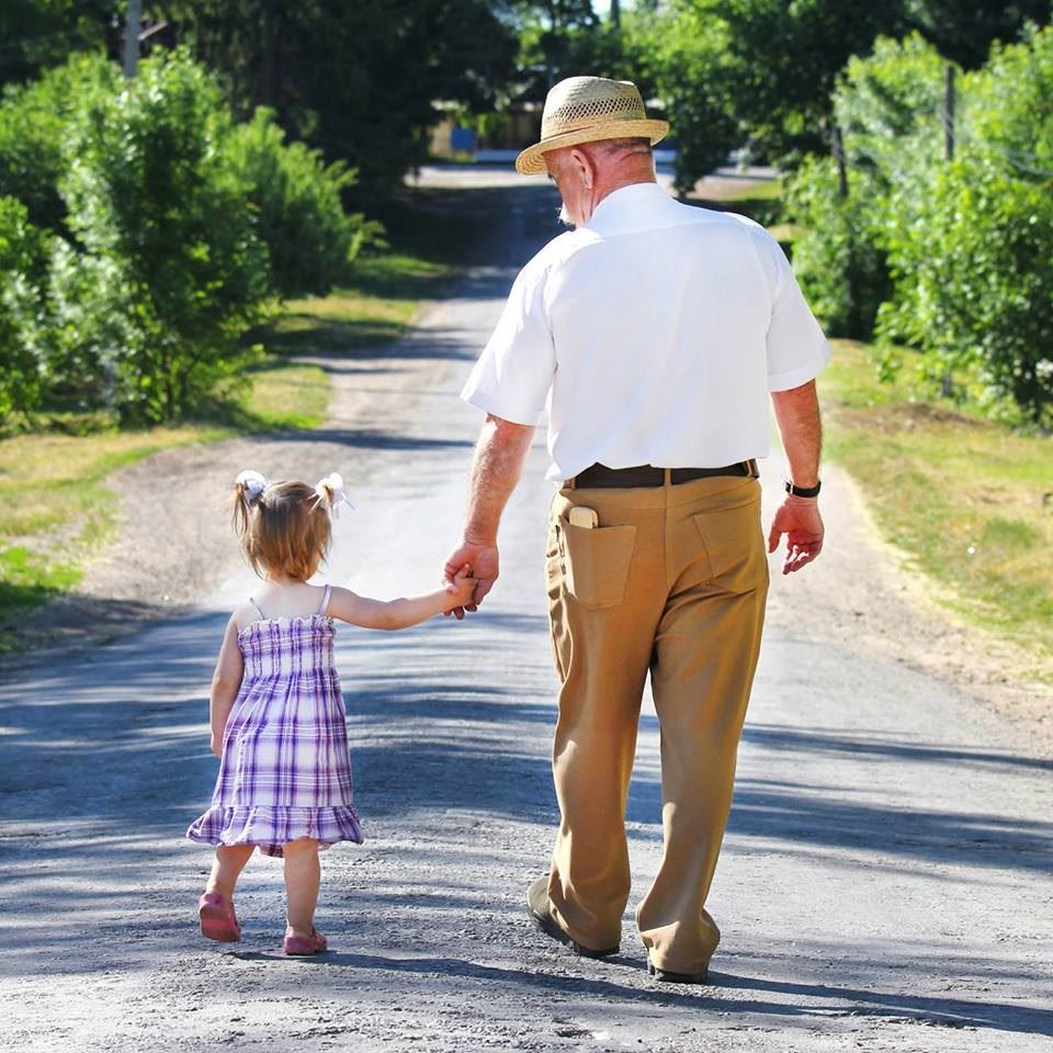 An older man in a hat walks down a back road hand-in-hand with a young girl.
